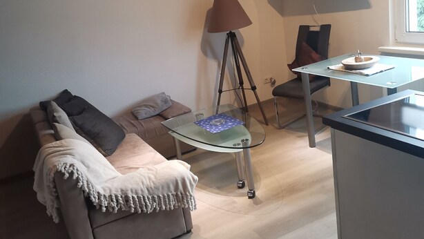 Ein modernes Wohnzimmer mit einem Glastisch, einer Couch mit Überwurfdecke, einer Stehlampe und einem flachbildfernseher an der Wand.
