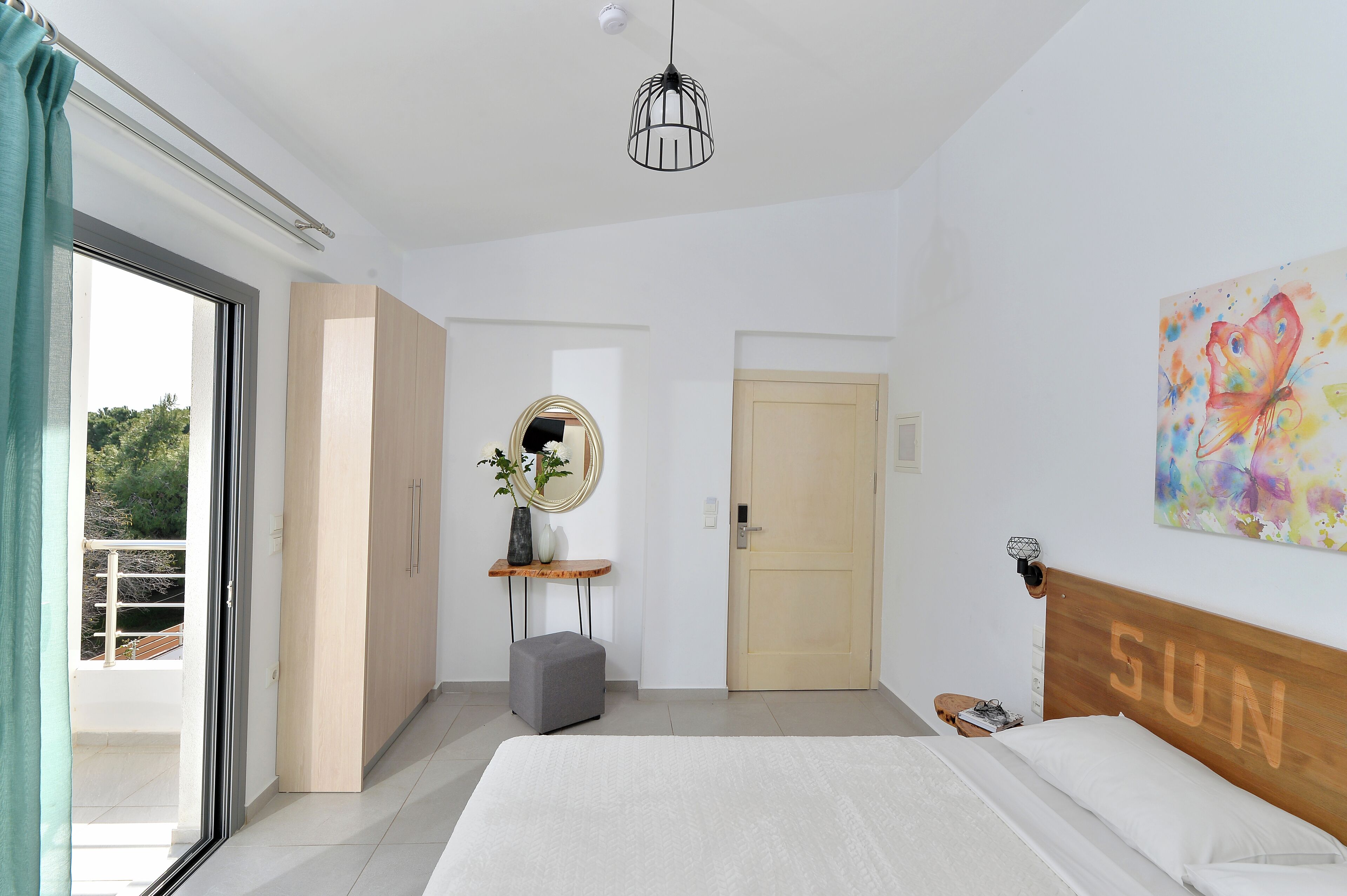 A bedroom with a bed, a bedside table, a nightstand, a mirror, a door, and a window with curtains.