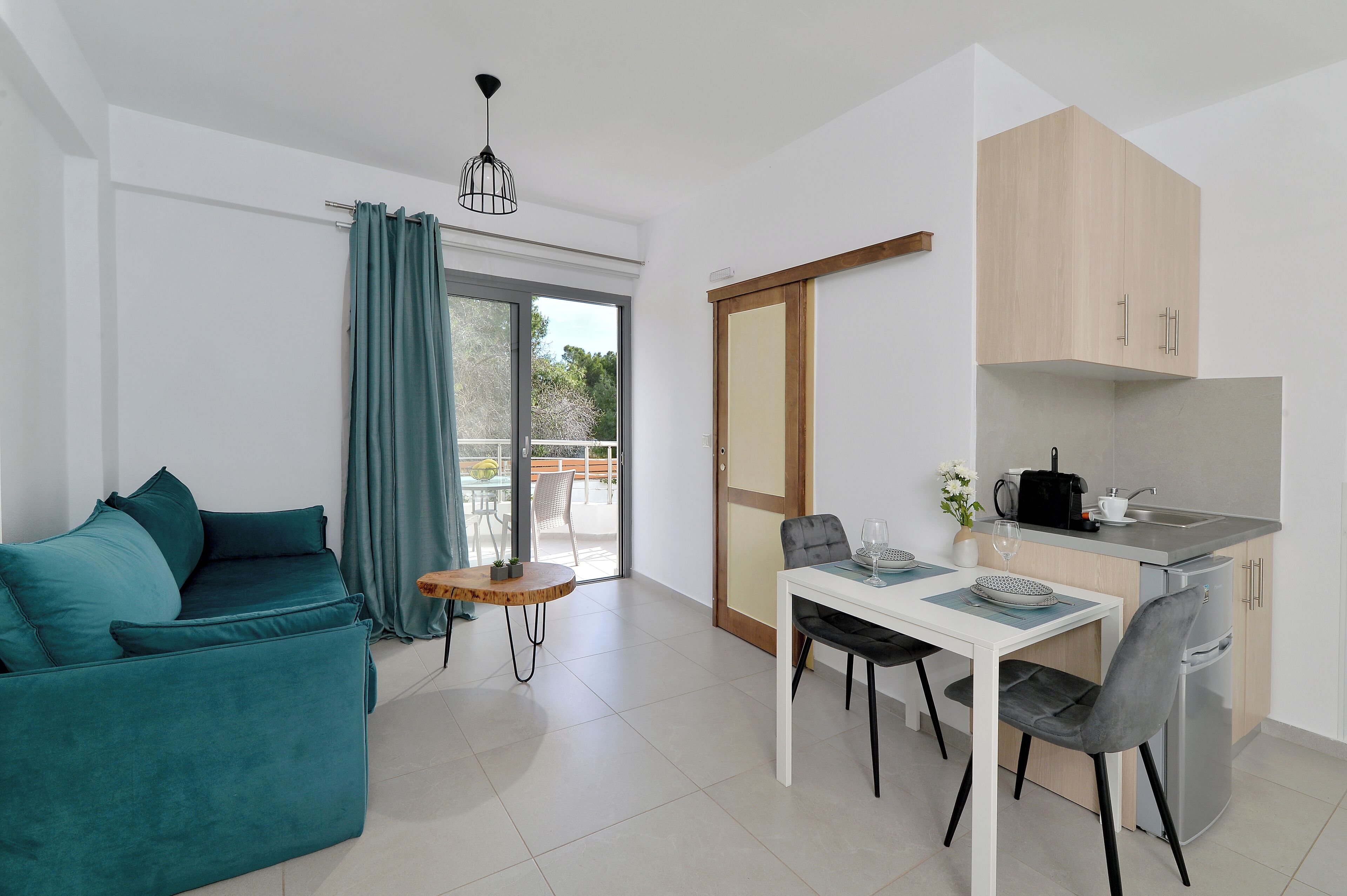Una sala de estar moderna con un sofá azul turquesa, una mesa de comedor con sillas, una zona de cocina con armarios, y un ventanal corredizo que da a un balcón.