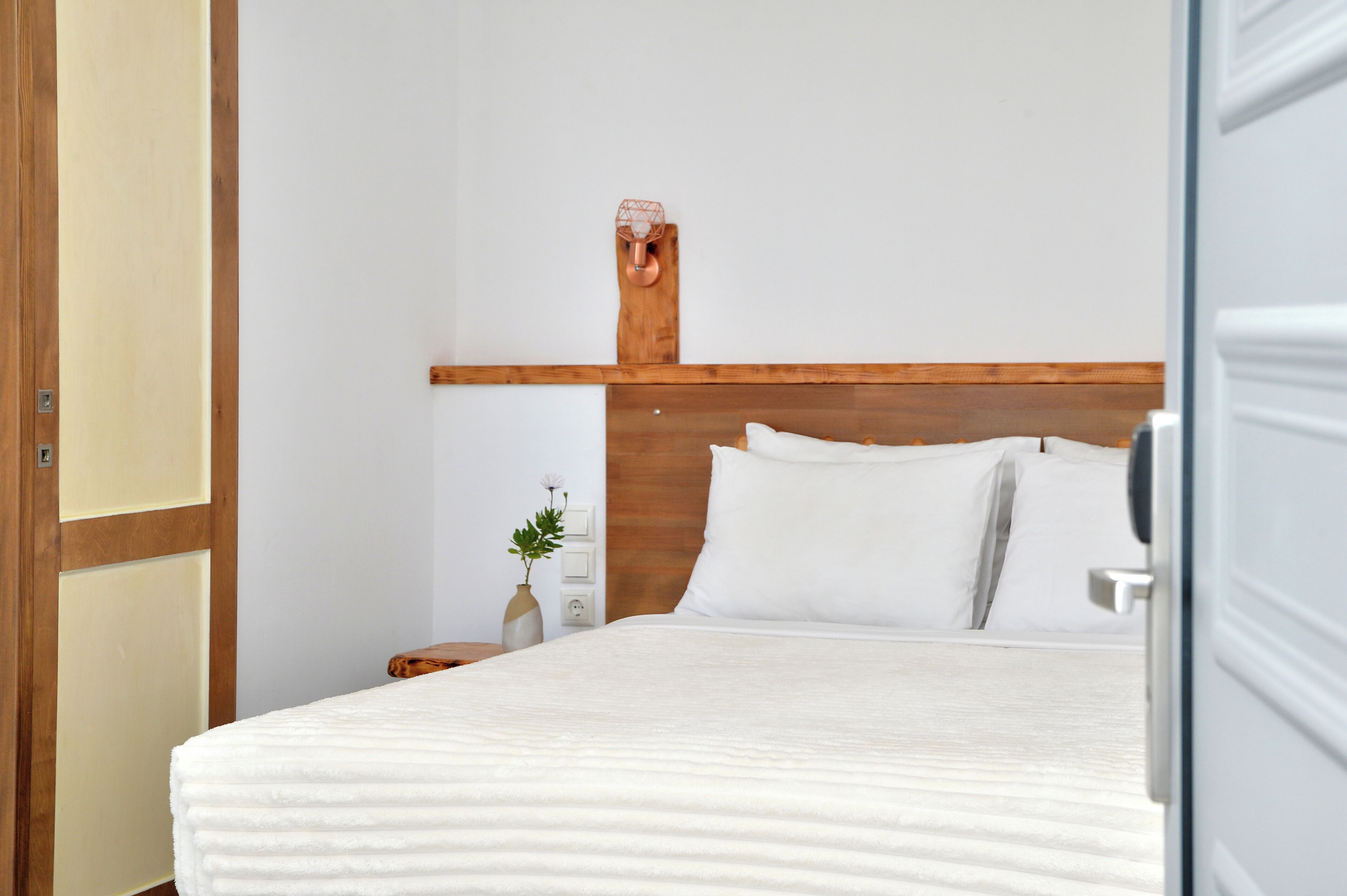 A neatly made bed with white linens in a room with wooden headboard and a simple nightstand with a vase of flowers.