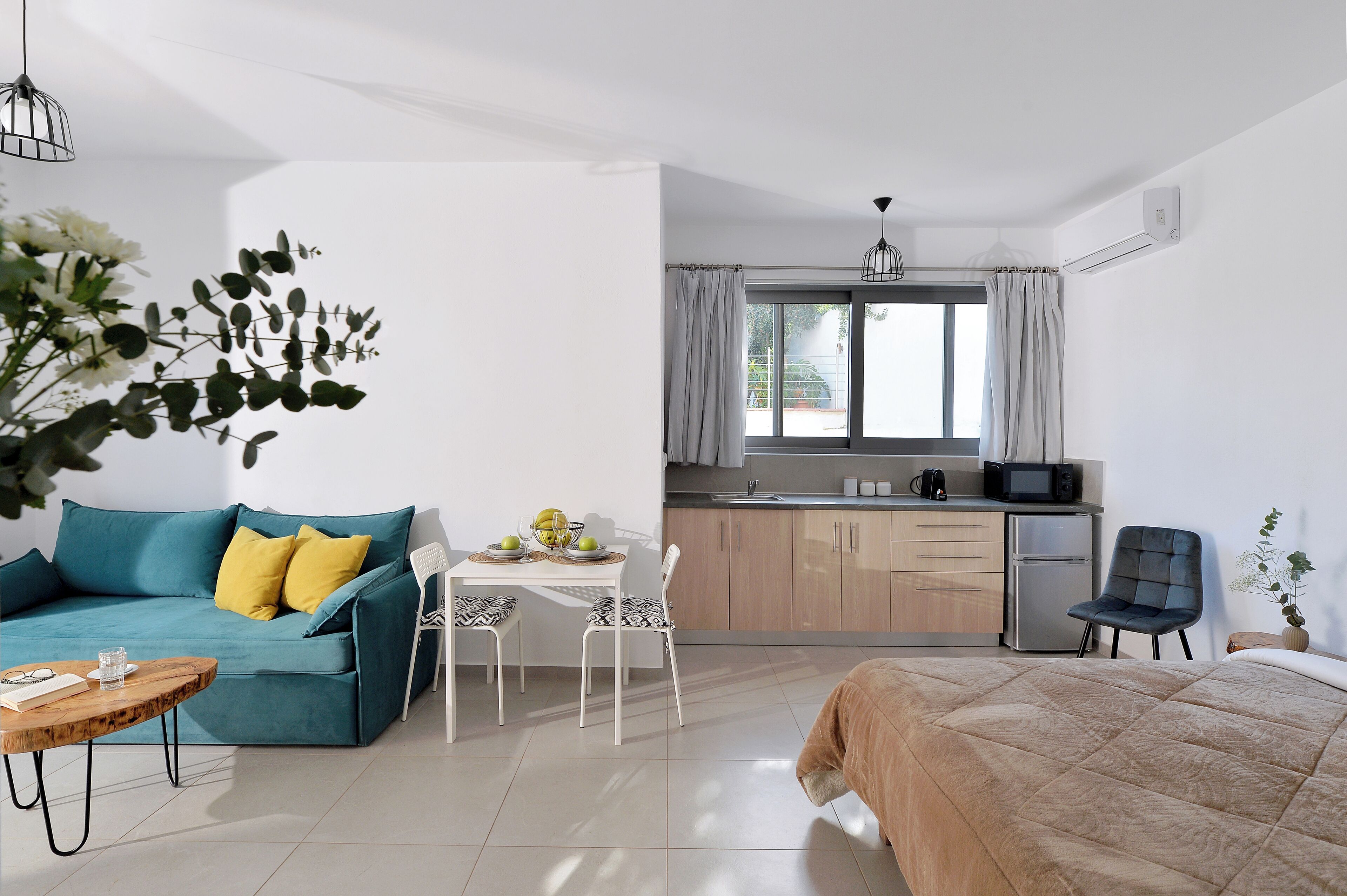 A modern room with a teal sofa, a wooden coffee table, a bed with a beige quilt, a white dining table with chairs, a kitchen area with a microwave, and a window with curtains.
