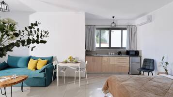 A modern room with a teal sofa, a wooden coffee table, a bed with a beige quilt, a white dining table with chairs, a kitchen area with a microwave, and a window with curtains.