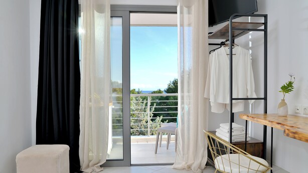 Una habitación moderna con puerta de vidrio corredera que da a un balcón, una pequeña mesa con una silla y un perchero con ropa colgada.