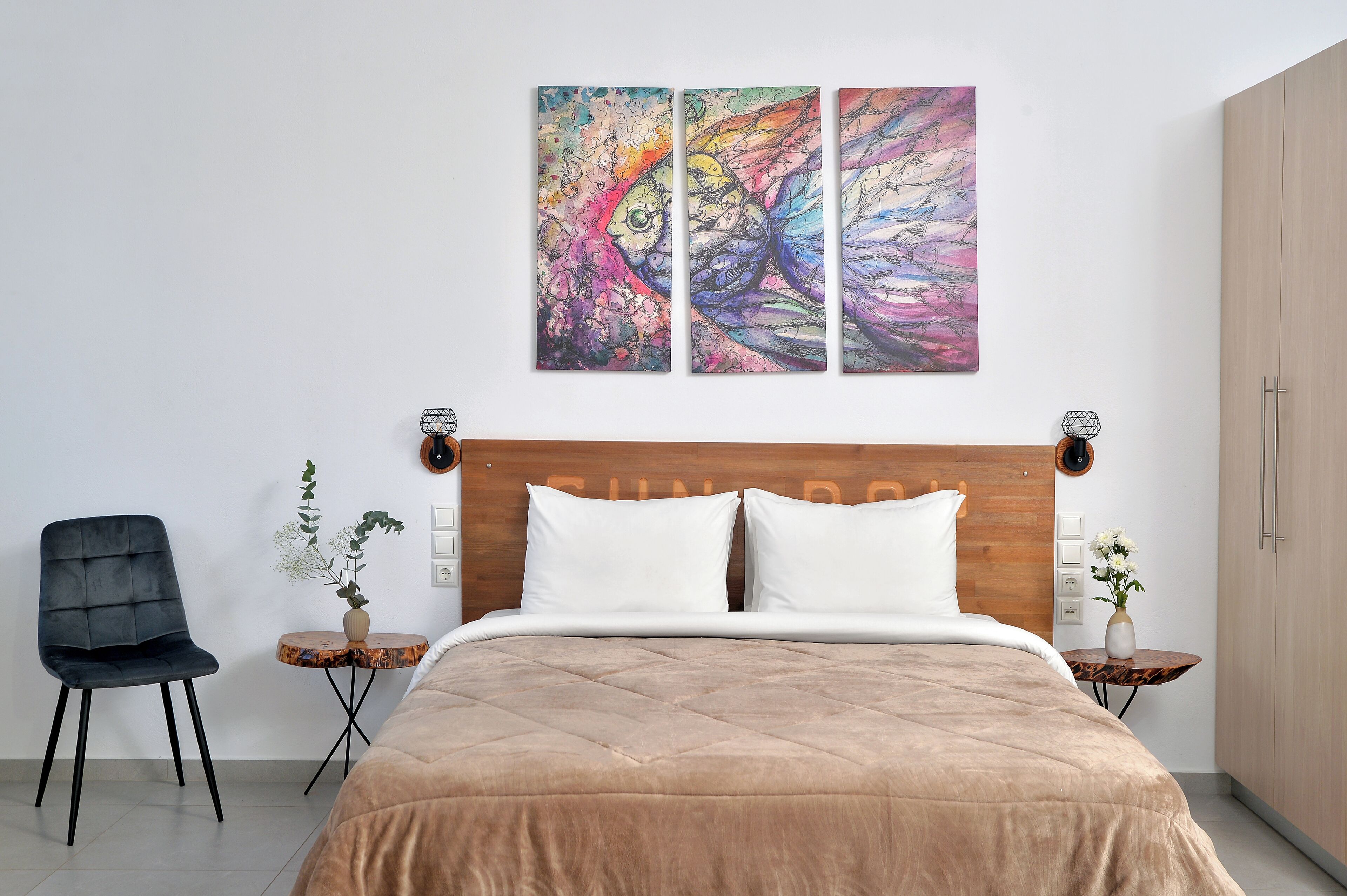 A modern bedroom with a large bed, a wooden headboard, a black chair, two bedside tables with vases, and a colorful abstract painting above the bed.