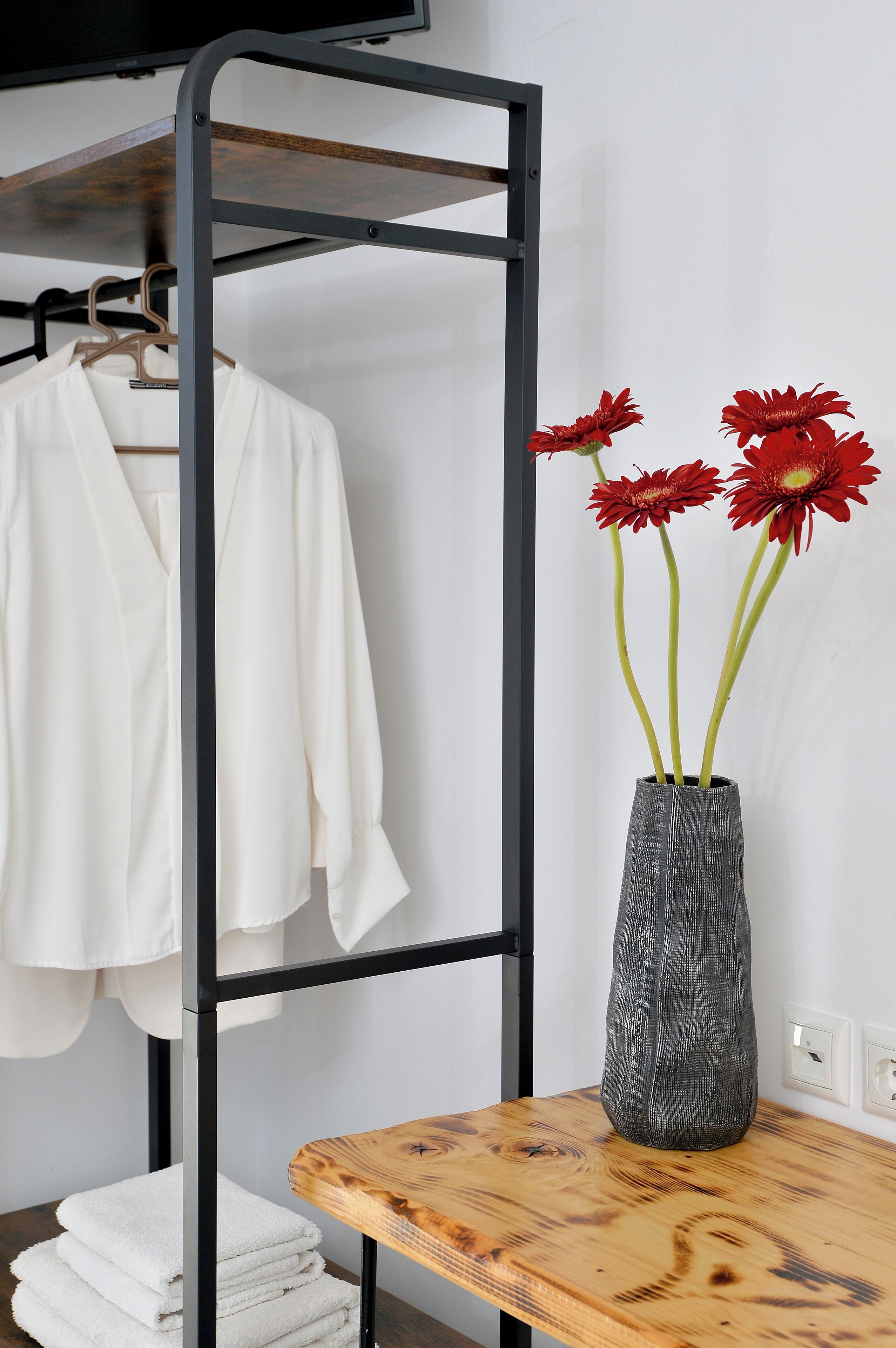 Una habitación con una estructura metálica que sostiene una camisa blanca, un jarrón gris con flores rojas y una mesa de madera con toallas blancas dobladas.