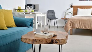 Una habitación de hotel moderna con un sofá azul, una mesa de centro de madera con un libro y vasos, una cama con colcha color beige, una pequeña mesa blanca con un cuenco de fruta y copas de vino, y una silla gris.