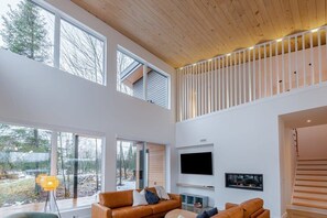 A modern interior with a high ceiling, large windows, a flat-screen TV, and a staircase.
