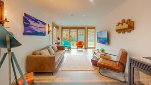 A modern living room with a sofa, armchairs, a coffee table, and a fireplace. There is a large window with a view of the outdoors.