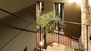A multi-story building with a modern design, featuring balconies with metal railings and a small garden area with plants.