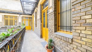 Un pasillo con puertas y ventanas amarillas, paredes de ladrillo y una barandilla de metal con plantas.