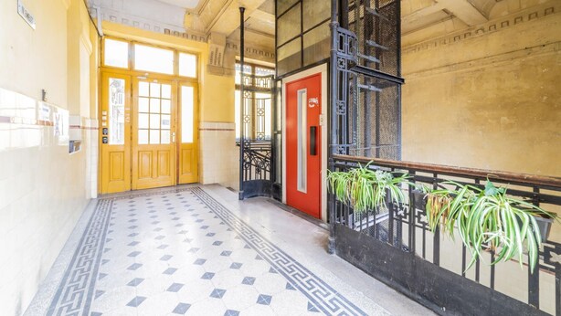 Un pasillo con piso estampado, una puerta roja y plantas en macetas en un balcón.