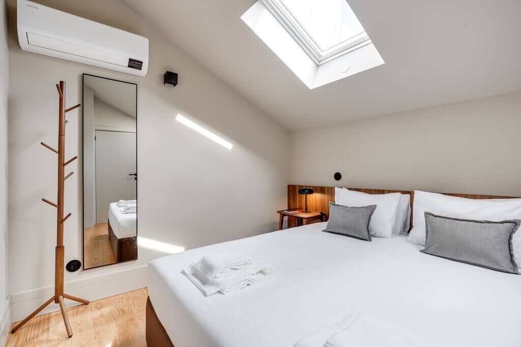 A hotel room with a skylight, a bed with white linens, a wooden coat rack, and a mirror.