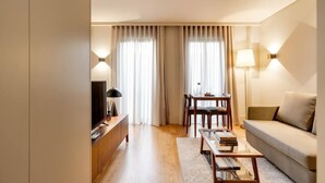 Habitació d'hotel moderna amb terra de fusta, un sofà, una taula de cafè, un televisor, un escriptori amb una làmpada i finestres grans amb cortines.
