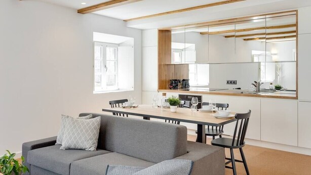 Un soggiorno moderno con un divano grigio, un tavolo da pranzo con sedie e un angolo cottura con travi in legno e pensili bianchi.