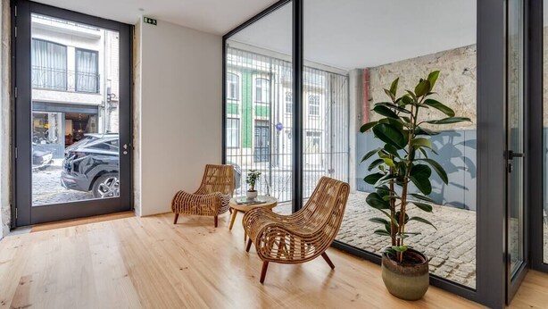 Una habitación moderna con ventanas amplias, sillas de mimbre, una mesita pequeña y una planta en maceta.