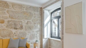 Una habitación con pared de piedra, un sofá amarillo con cojines grises, un taburete de madera y una ventana con vista a edificios.