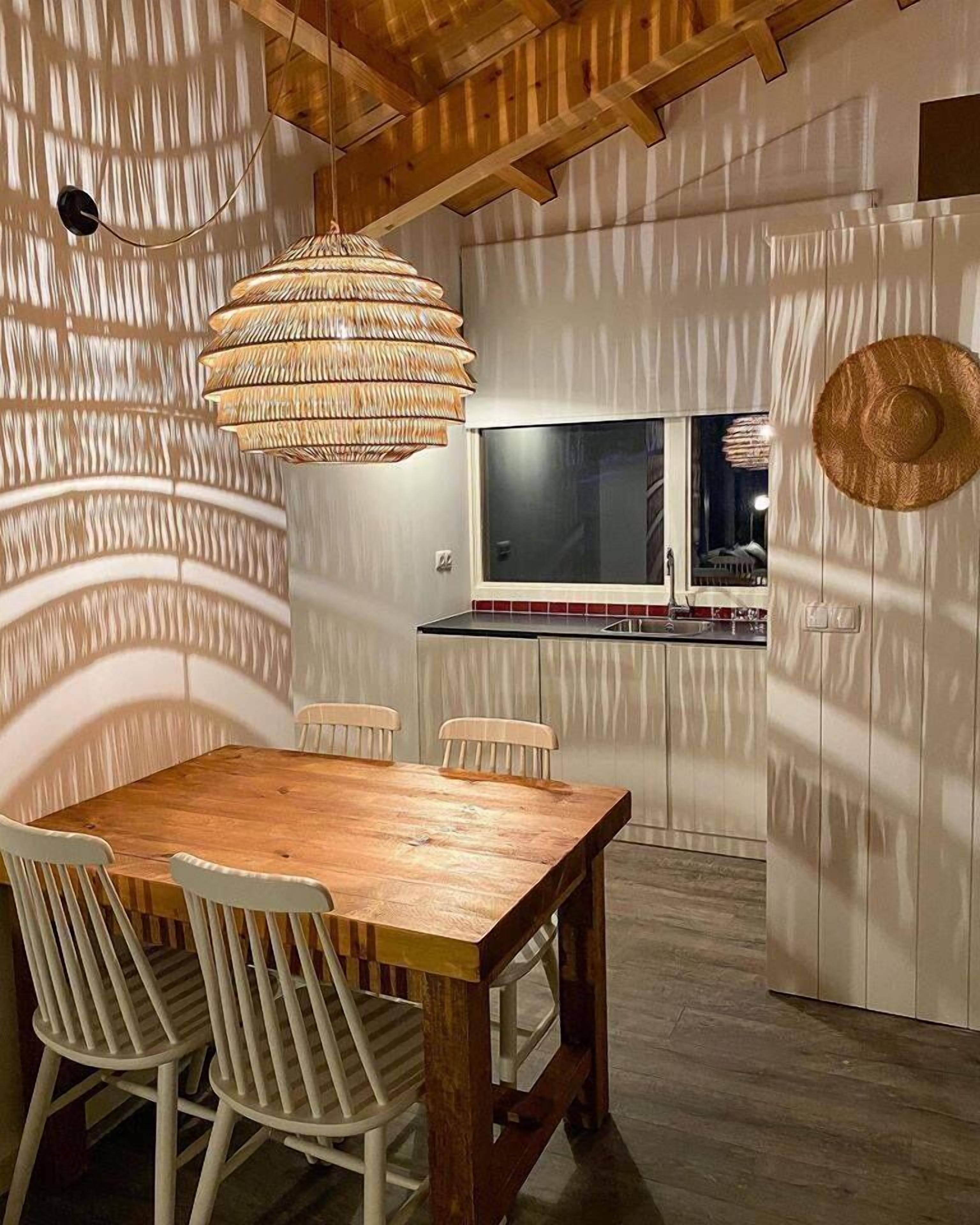 A dining area with a wooden table and chairs, a woven pendant light, and a view of the kitchen through a window.