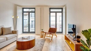 Ein modernes Wohnzimmer mit einer Couch, einem runden Couchtisch, einem Holzstuhl, einem Fernseher und einem großen Fenster mit Blick auf Gebäude.