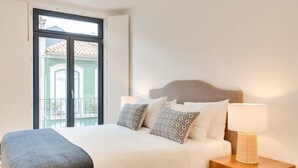 Ein Hotelzimmer mit einem Bett, Kissen, einem Nachttisch mit Lampe und einem Balkon mit Blick auf Gebäude.