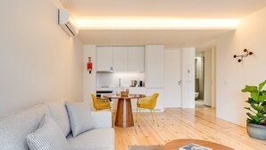 Een moderne woonkamer met een grijze bank, een houten salontafel en gele stoelen. In de achtergrond is een keuken met witte kasten en een eettafel zichtbaar.