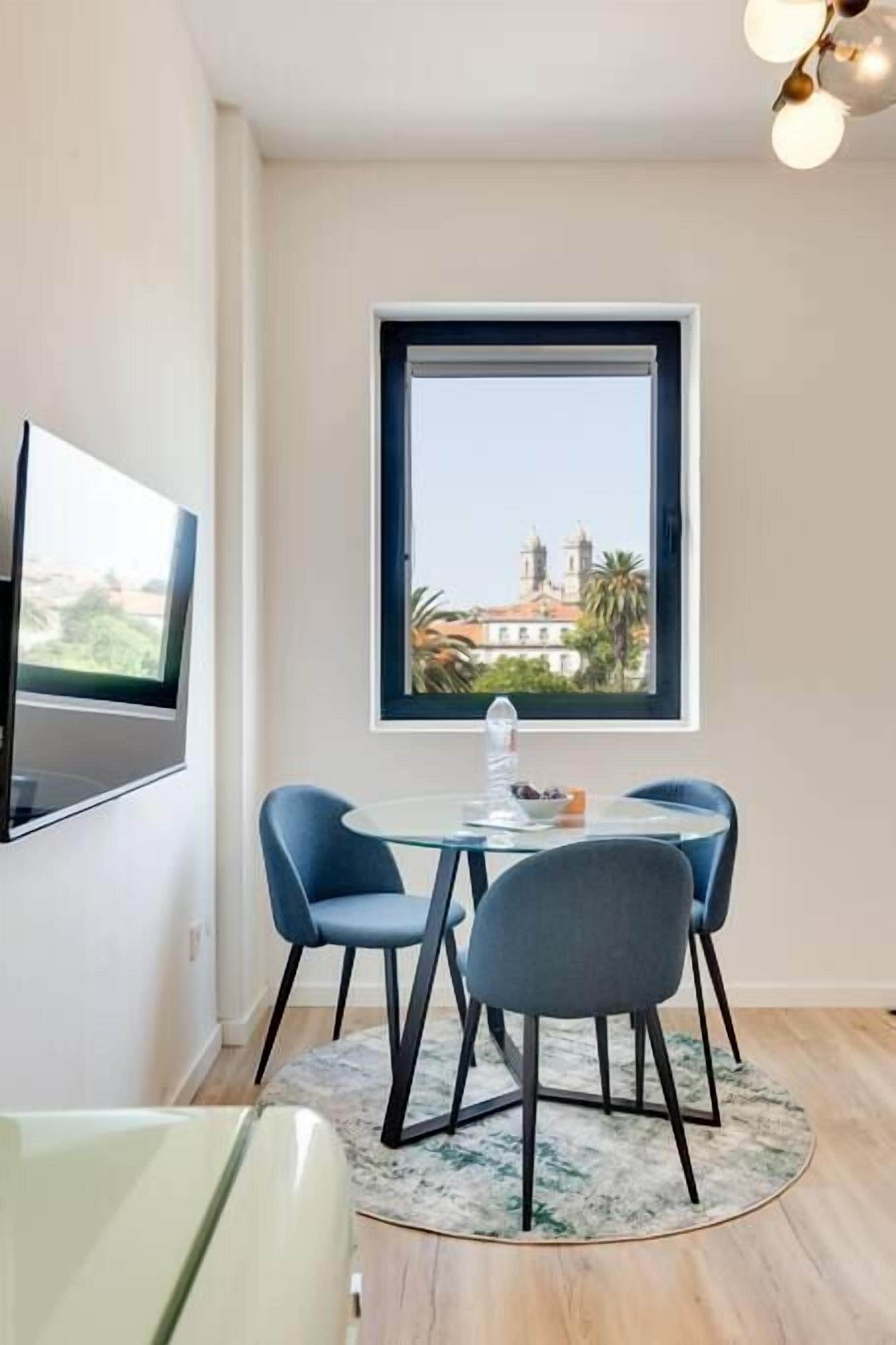 Une chambre moderne équipée d’une table à manger, de deux chaises, d’un téléviseur fixé au mur et d’une fenêtre donnant sur le paysage urbain.