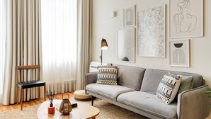 Ein Wohnzimmer mit grauem Sofa, einem Holzstuhl, einem runden Couchtisch und einer Lampe. An der Wand befinden sich gerahmte Kunstwerke.