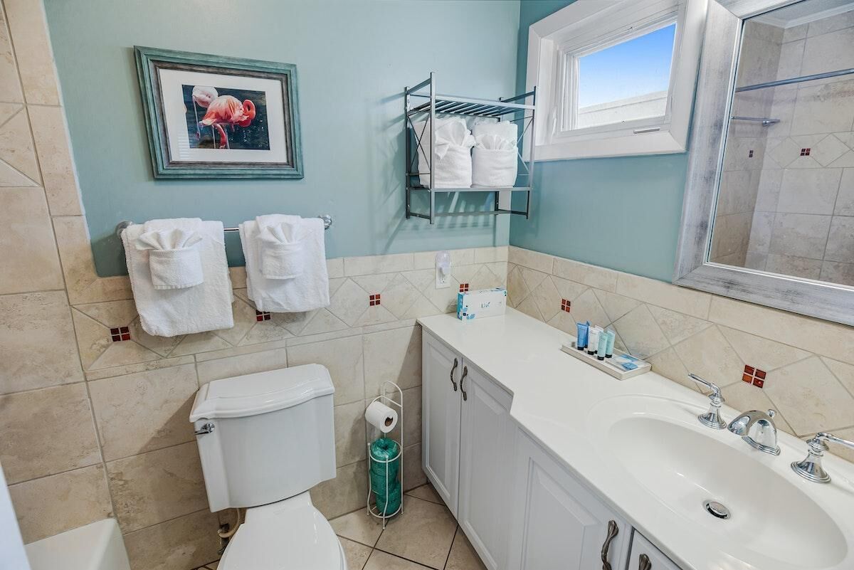 Un baño con inodoro, lavamanos y zona de ducha. Hay toallas colgadas en un perchero, un cuadro en la pared y una ventana que proporciona luz natural.