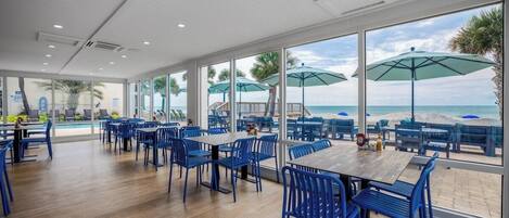 A spacious indoor dining area with large windows overlooking a beach, wooden flooring, and blue chairs.