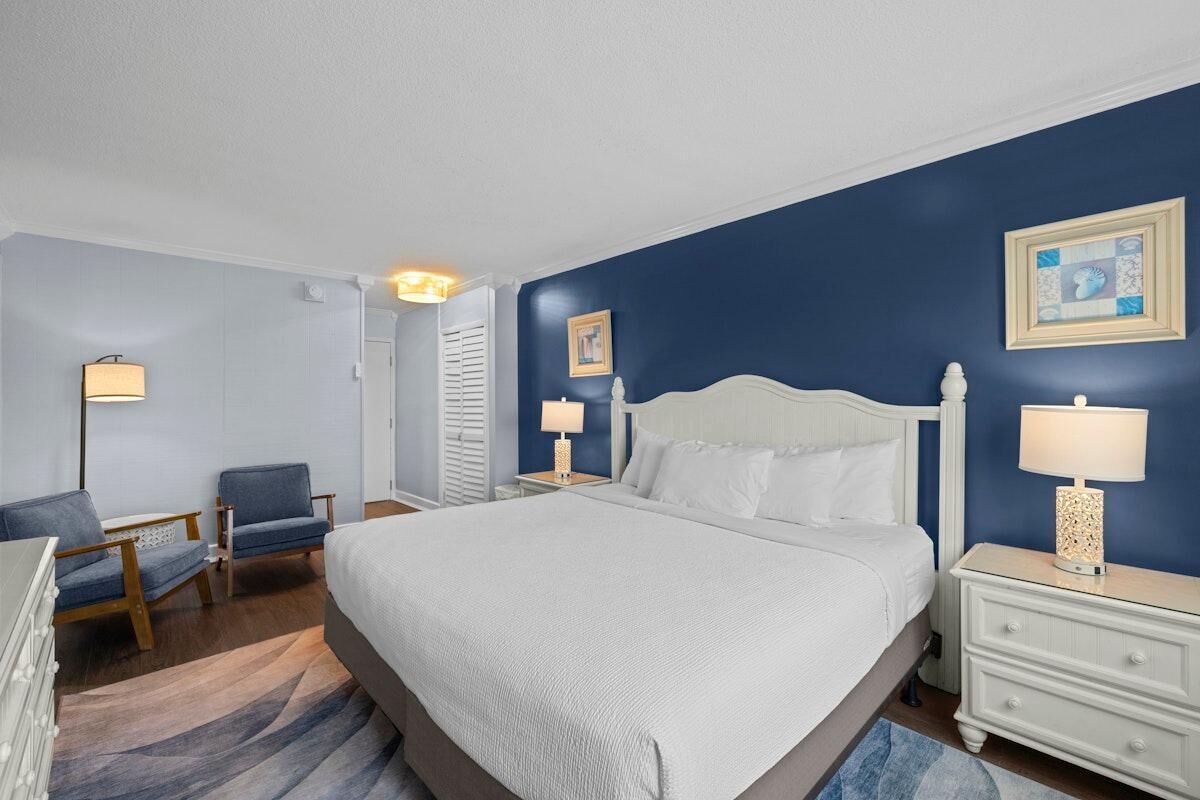 Une chambre d’hôtel avec un grand lit, deux tables de chevet avec des lampes, une chaise, une commode et un mur d’accent bleu.