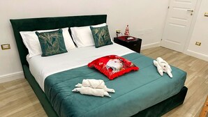 A neatly made bed with a teal bedspread, white pillows, and a red decorative pillow. There are two white towels on the bed and a small nightstand with a red and white decorative item. - Quadruple Room. Berenice Suites (Napoli)