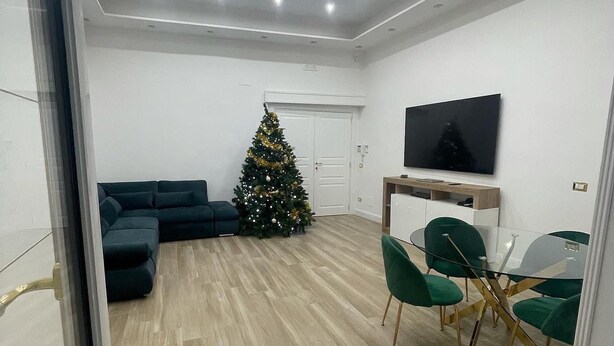 A modern living room with a wooden floor, a green sofa, a glass dining table with green chairs, a Christmas tree, and a flat-screen TV mounted on the wall. - Quadruple Room. Berenice Suites (Napoli)