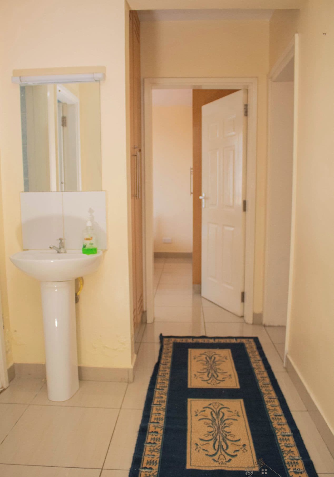Un couloir avec un lavabo et un miroir blancs, un tapis à motifs et une porte donnant sur une autre pièce.
