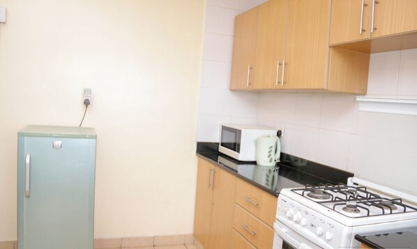 A compact kitchen with a small refrigerator, a microwave, a stove, and wooden cabinets. - Lux Suites Tulia Homestays Nakuru (Nakuru)