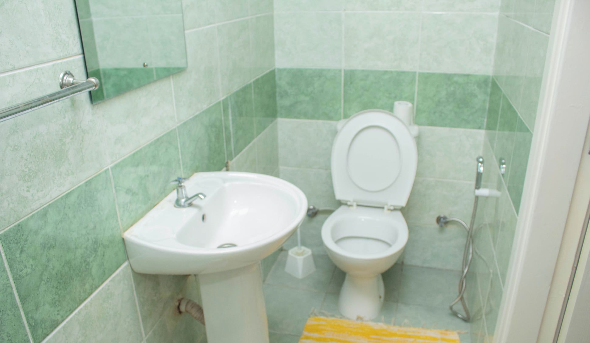 Et badeværelse med vask, toilet, grønne fliser, brusebad og en gulstribet måtte.