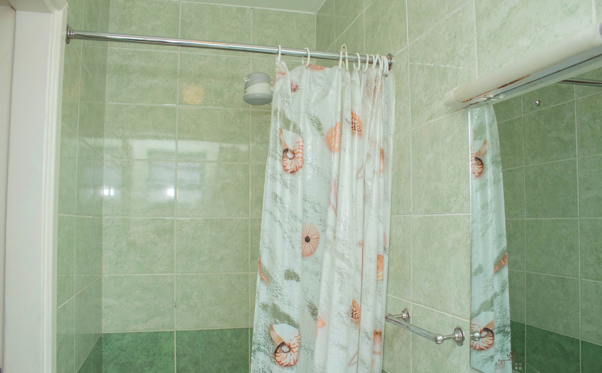 A bathroom with green tiles, a shower curtain with a shell pattern, and a metal grab bar.