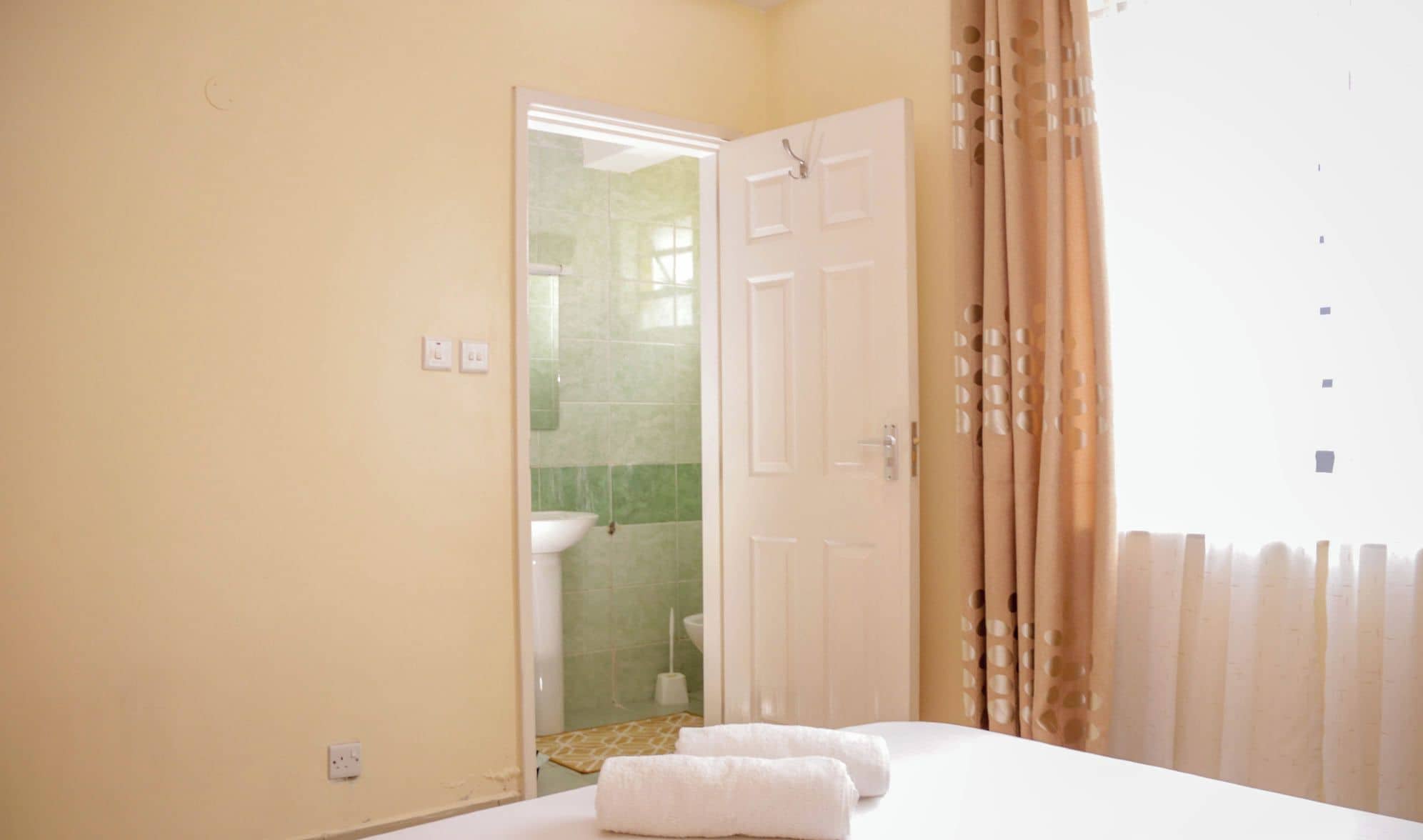 Une chambre d’hôtel avec un lit, deux serviettes, une porte donnant sur une salle de bain et une fenêtre avec des rideaux.