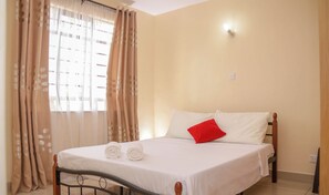 A neatly made bed with white linens and a red pillow, a window with beige curtains, and a light switch on the wall. - Lux Suites Tulia Homestays Nakuru (Nakuru)