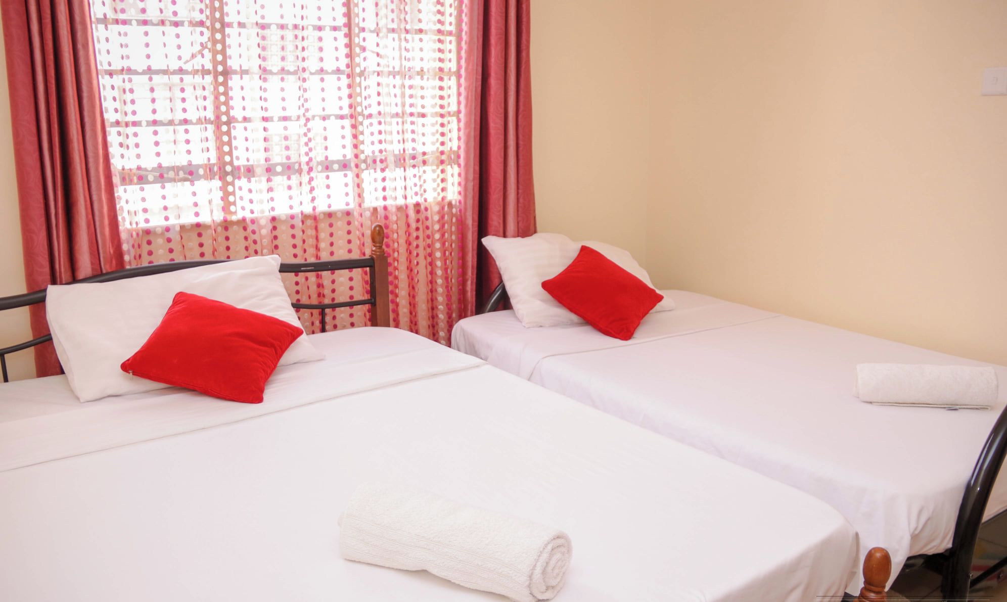 A hotel room with two single beds, each with white bedding and red pillows. A rolled white towel is placed on the floor beside one bed.