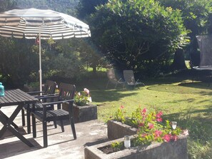 Une terrasse avec une table et des chaises, un parasol à rayures et un petit jardin fleuri.
