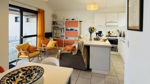 A modern living room with a sofa, chairs, and a dining table. There is a kitchen area with a stove, sink, and cabinets. A bookshelf is also visible. - 3-Room Apartment for 6, Steps from Beach, Terrace, Parking Included (Lumio)