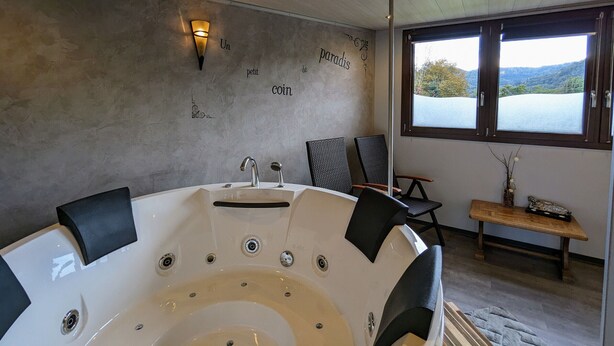 A modern bathroom with a freestanding bathtub, a wall-mounted light fixture, and a view of a lake through a window. - Charming Roulotte Stay: Streamside Serenity, Sauna, Massage & Ideal for Family or Friends (Ferdrupt)