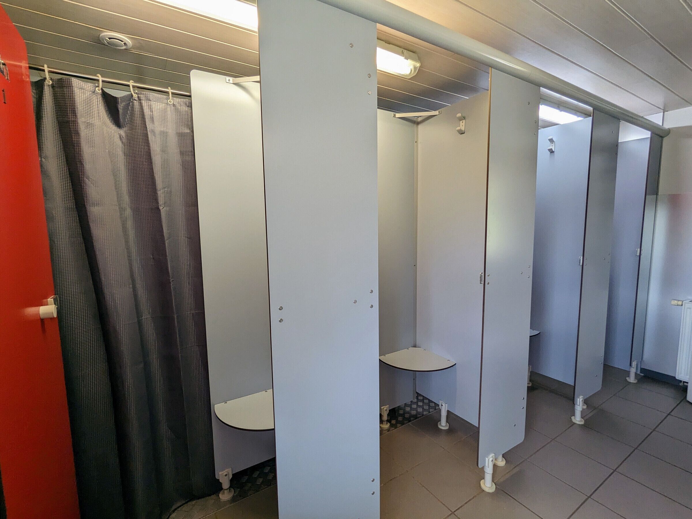 Des toilettes publiques avec plusieurs cabines, un espace douche et une porte rouge.