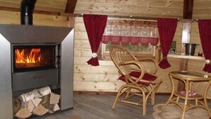 A cozy cabin interior with a wood-burning stove, wicker chairs, a small table, and a window with curtains.