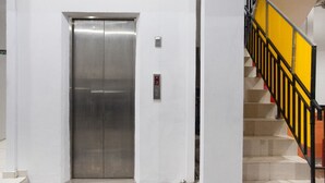 Un ascenseur aux finitions métalliques, un escalier avec des rampes jaunes et noires, et un panneau de commande fixé au mur.
