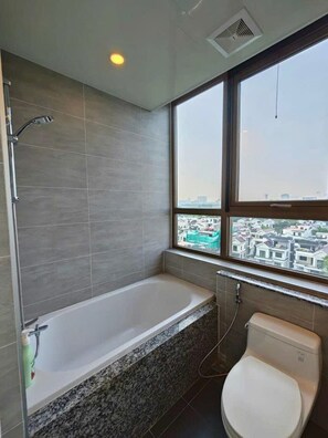 A modern bathroom with a bathtub, toilet, and a window offering a city view. - Starlake Daewoo Managed by Anstay Ha Noi (Hanoi)