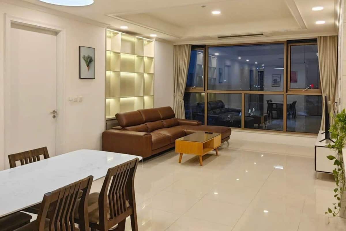 A modern living room with a dining area, featuring a large window, a brown leather sofa, a wooden coffee table, and a white dining table.