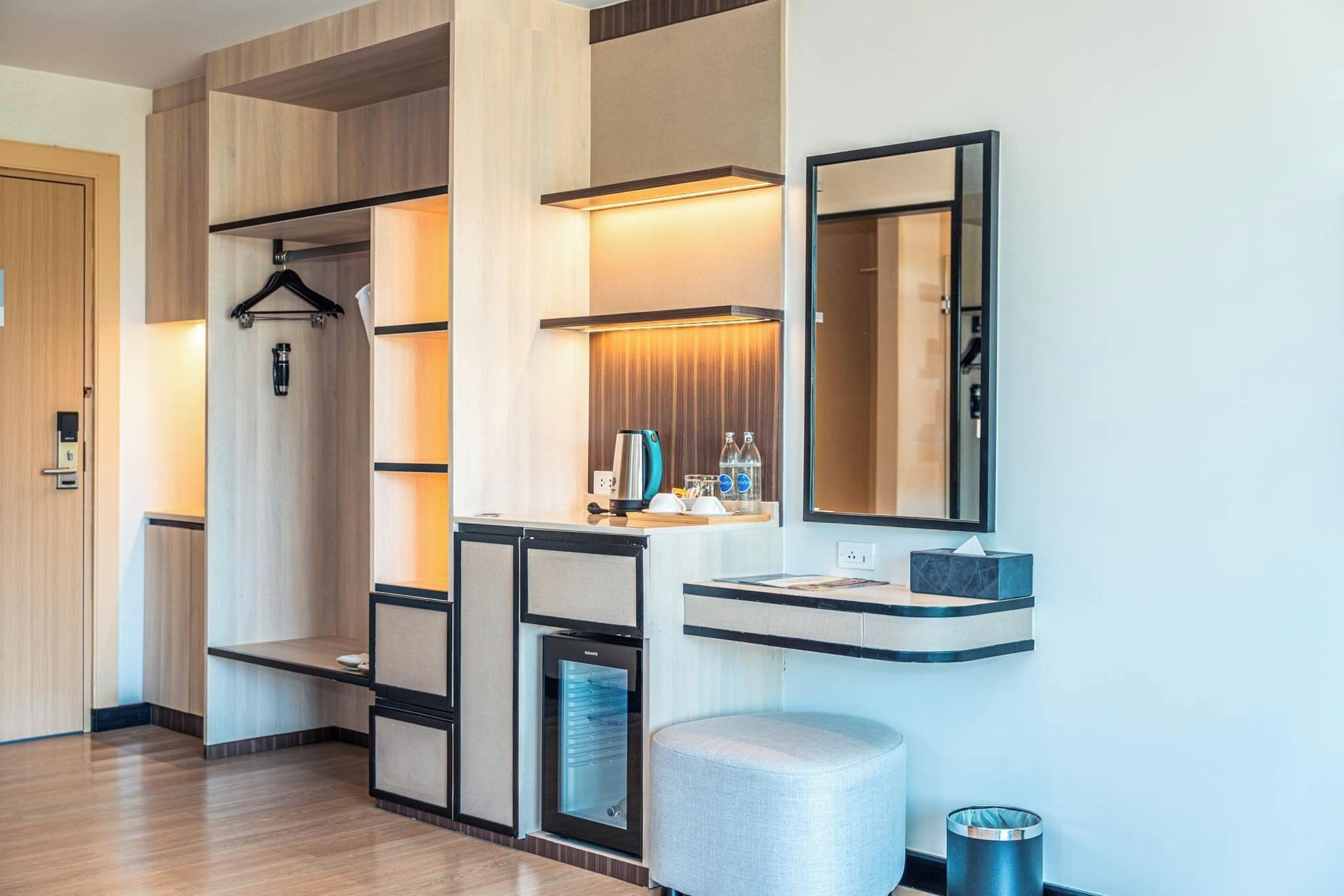 Una habitación de hotel moderna con suelo de madera, armario empotrado, minibar, refrigerador pequeño, espejo y cesto de basura.