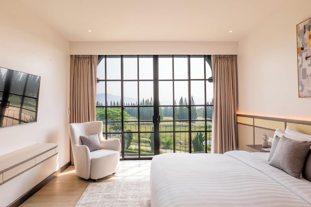 A bedroom with a large window, a bed, a chair, a TV, and a bedside table.