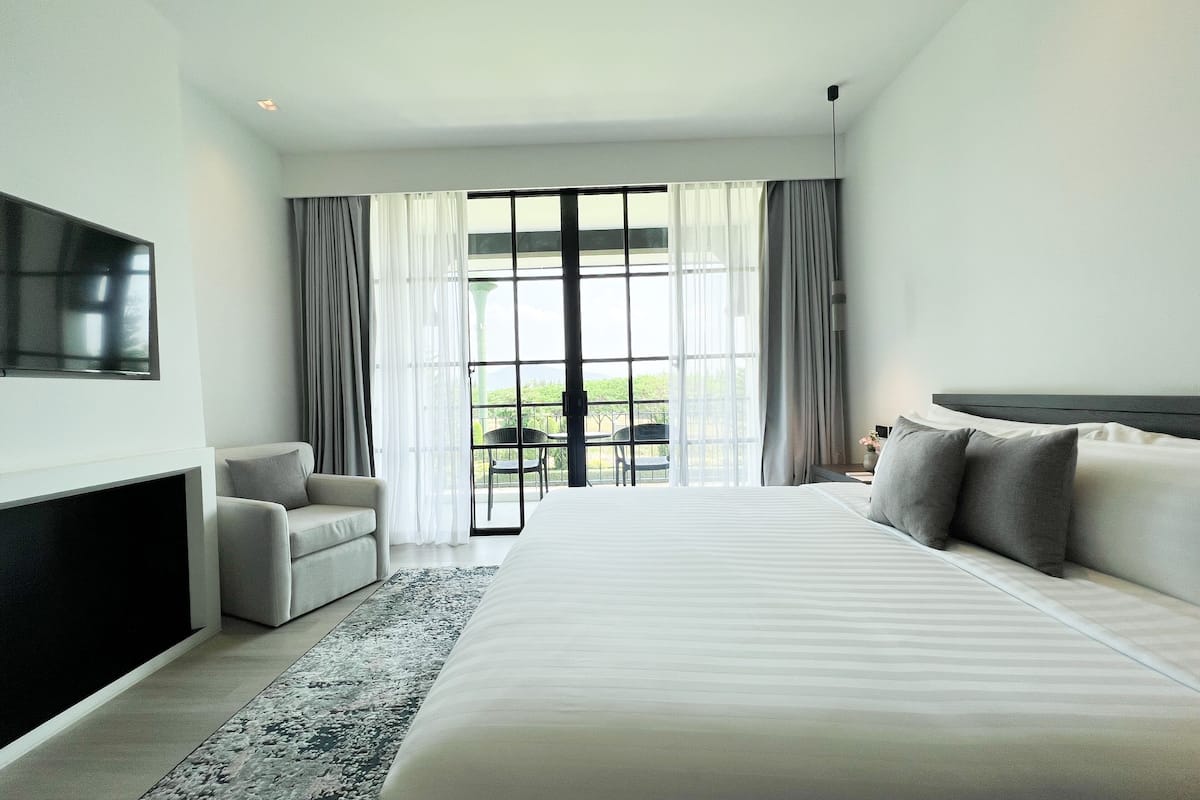 A modern hotel room with a large bed, a flat-screen TV mounted on the wall, a seating area with a sofa and armchair, and a balcony with a view.