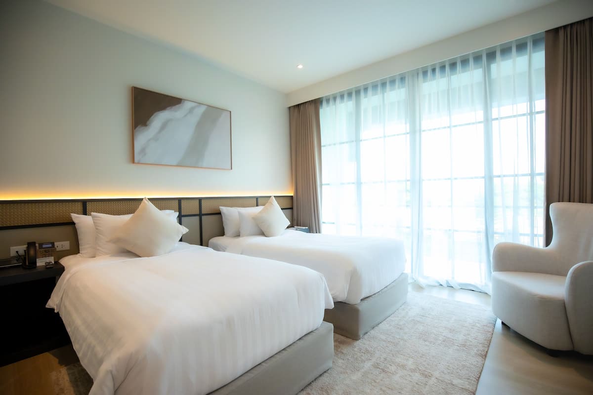 A hotel room with two beds, a sitting area, a painting on the wall, and large windows with curtains.